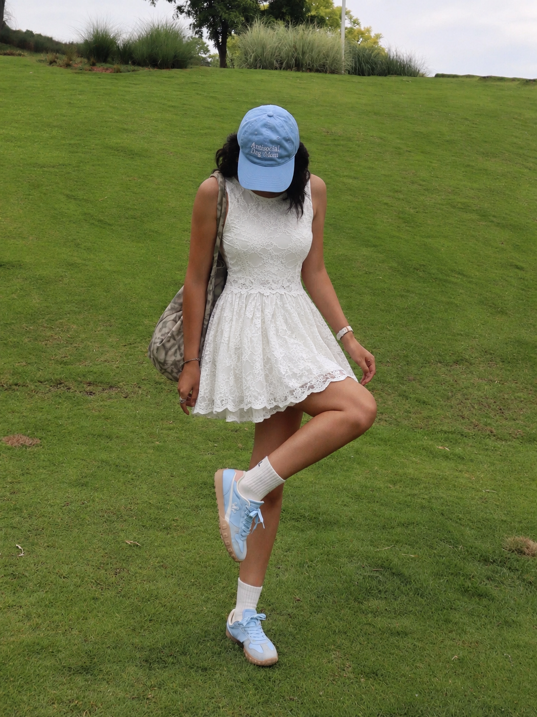 White lace dress styled with a pale blue cap, blue sneakers, white crew socks, and a shoulder bag on a lawn.