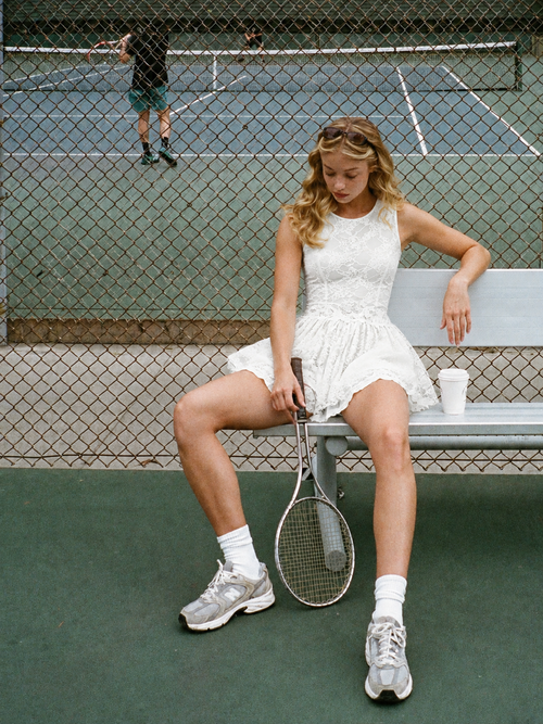 Woman in Crayon talk Grace Lace Tennis Dress sitting courtside on a bench, holding a racket